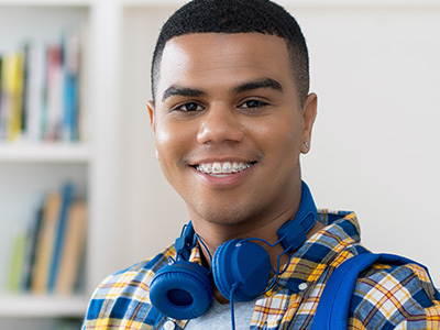 The image shows a young man with a bright smile, wearing headphones and a plaid shirt, standing in front of a bookshelf.