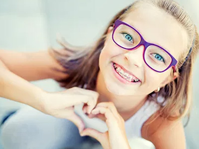 A young girl with glasses and a heart-shaped sign is making a heart gesture in front of her face.