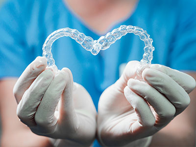A dental hygienist holding a transparent heart-shaped object, possibly a dental appliance or model, against a background that suggests a medical setting.