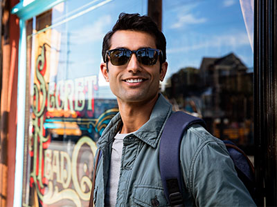 The image is a portrait of a man standing in front of a storefront, wearing sunglasses and a backpack. He has a beard and mustache, and appears to be posing for the photograph.