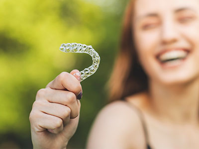 The image shows a person holding up a clear plastic tray with the word  SMILE  written on it, which is likely meant to convey the idea of dental braces or aligners.