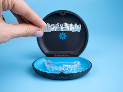 The image shows a hand holding an open container displaying a clear dental retainer with a blue lid.